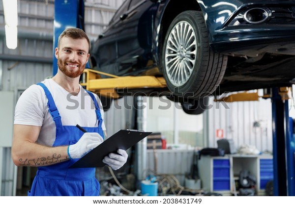 Young happy technician mechanic man wears denim\
overalls use hold clipboard papers document writing estimate stand\
near car lift check technical condition work in vehicle repair shop\
workshop indoors