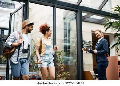 Young Happy Tourist Couple Arriving At The Hotel While Female Receptionist Welcoming Them At The Entrance. 