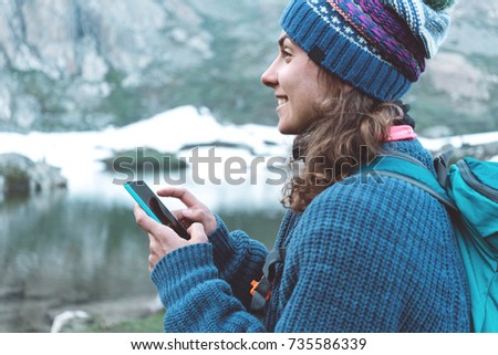 Young happy traveler girl with backpack, hat, sweater typing on mobile phone, smiling in the stunning mountain wilderness in front of amazing cold lake after day of hiking.