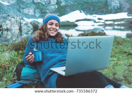 Young happy traveling girl with backpack, hat lying in the grass and flowers and using laptop, smiling, drinking hot tea in the stunning mountain wilderness after day of hiking.