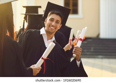 Young happy university graduate in a mantle demonstrates his diploma and smiles at the camera next to his classmates. Concept of success and goal achievement. - Powered by Shutterstock