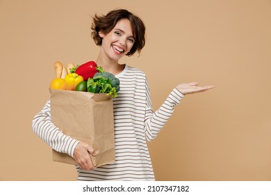 Young Happy Vegetarian Woman 20s In Casual Clothes Hold Paper Bag With Vegetables Point Hand Arm Aside On Workspace Area Copy Space Mock Up Isolated On Plain Pastel Beige Background Shopping Concept