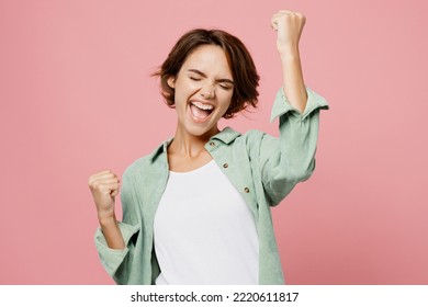 Young happy woman 20s she wear green shirt white t-shirt doing winner gesture celebrate clenching fists say yes isolated on plain pastel light pink background studio portrait. People lifestyle concept