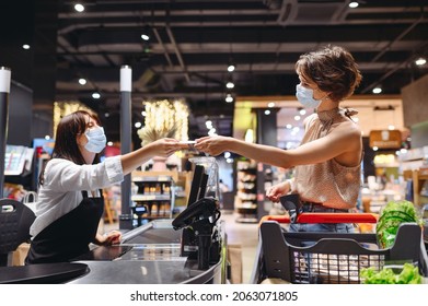 Young happy woman 20s wearing casual clothes mask shopping at supermaket stand at store checkout giving money to cashier pay inside hypermarket. People lifestyle purchasing gastronomy food concept.