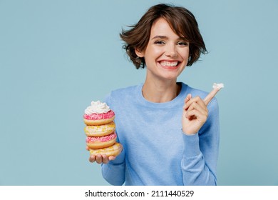 Young happy woman 20s wearing casual sweater look camera hold donuts dessert show finger with cream isolated on plain pastel light blue background studio. People lifestyle junk unhealthy food concept