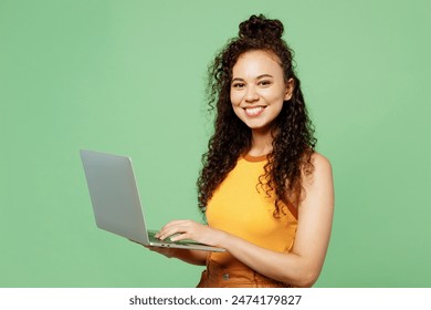 Young happy IT woman of African American ethnicity wears yellow tank shirt top hold use work on laptop pc computer isolated on plain pastel light green background studio portrait. Lifestyle concept