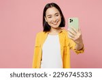 Young happy woman of Asian ethnicity wear yellow shirt white t-shirt doing selfie shot on mobile cell phone post photo on social network isolated on plain pastel light pink background studio portrait