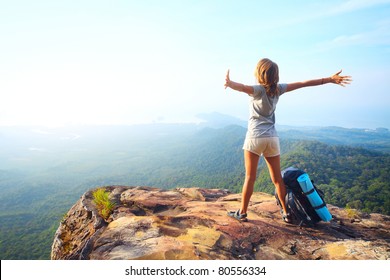 Young happy woman with backpack standing on a rock with raised hands and looking to a valley below