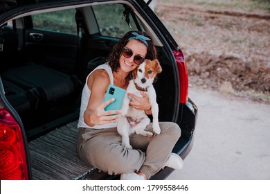 Young Happy Woman In A Car Enjoying With Her Cute Dog. Taking A Selfie With Mobile Phone. Travel Concept