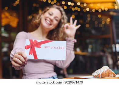 Young Happy Woman In Casual Clothes Sit At Cafe Table Eat Breakfast Show Ok Gesture Hold Gift Certificate Coupon Voucher Card For Store Relaxing In Restaurant During Free Time Indoors. Focus On Card