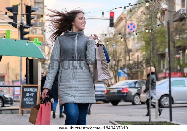 Young happy woman in coat with paper shopping bags\
walking along city street, season winter spring, seasonal discounts\
and sales