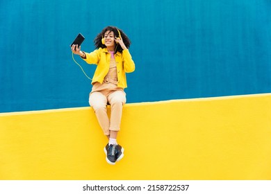 Young happy woman dancing and having fun outdoor. Teenager listening to music with smartphone and headphones in a yellow and blue modern urban area - Shutterstock ID 2158722537