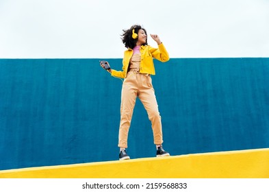 Young happy woman dancing and having fun outdoor. Teenager listening to music with smartphone and headphones in a yellow and blue modern urban area - Shutterstock ID 2159568833