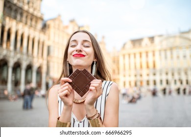 Young And Happy Woman With Dark Chocolate Bar Standing Outdoors On The Grand Place In Brussels In Belgium. Belgium Is Famous Of Its Chocolate