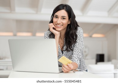 Young Happy Woman Doing Shopping Online With Her Laptop At Home. Portrait Of Smiling Woman In Casual Holding Credit Card And Looking At Camera. Beautiful Girl Paying Online Bills Using Gold Card.