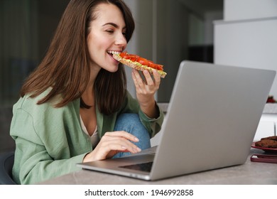 Young Happy Woman Eating Avocado Toast While Working With Laptop At Home Kitchen