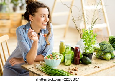 Young and happy woman eating healthy salad sitting on the table with green fresh ingredients indoors - Shutterstock ID 622381814
