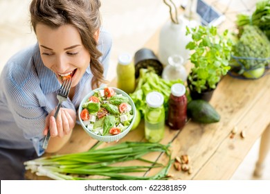 Young and happy woman eating healthy salad sitting on the table with green fresh ingredients indoors - Shutterstock ID 622381859