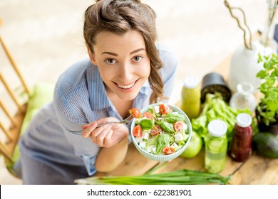 Young And Happy Woman Eating Healthy Salad Sitting On The Table With Green Fresh Ingredients Indoors