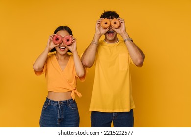 Young Happy Woman And Enthusiastic Man Holding Donuts On Eyes , While Standing Over Isolated Yellow Background