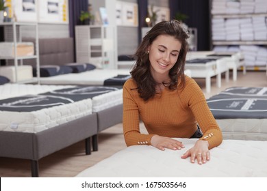 Young Happy Woman Examining Orthopedic Mattress On Sale At Furniture Store. Charming Female Customer Choosing New Bed To Buy