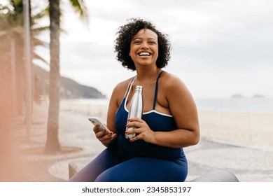 Young, happy woman in fitness clothing sits on a promenade by the beach, taking a break from her workout to use her smartphone. Woman engaging in a healthy exercise routine with a cheerful smile. - Powered by Shutterstock