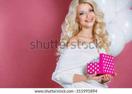 Young happy woman with a gift. Happy birthday. Sweet blonde woman holding small gift box with ribbon. Soft colors. Studio portrait over pink background. 