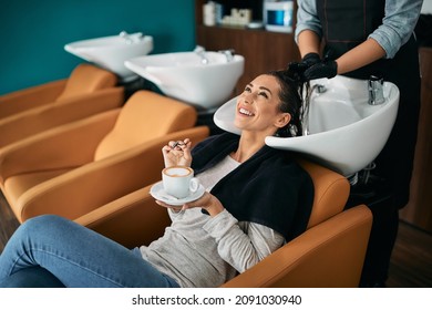 Young happy woman having cup of coffee while washing hair at hair salon.