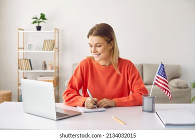Young happy woman at home learning English attending online courses using laptop. Smiling girl writes information on notebook while watching educational webinar sitting near american flag on table. - Shutterstock ID 2134032015