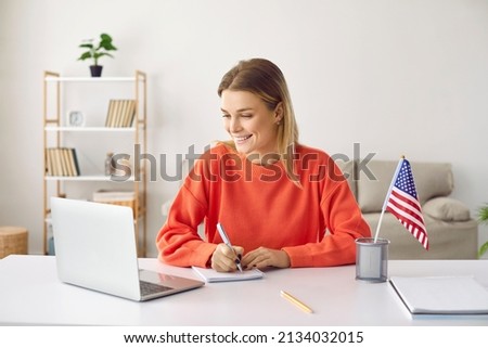Young happy woman at home learning English attending online courses using laptop. Smiling girl writes information on notebook while watching educational webinar sitting near american flag on table.