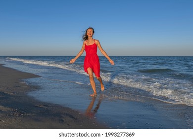 Young Happy Woman Jog On The Beach And Enjoy Life. Smiling Girl In Red Dress Run At Sea Shore Water