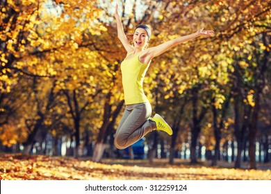 Young Happy Woman Jumping In Autumn Park. Female Fitness Model Training Outside On A Warm Fall Day. Sport Lifestyle.