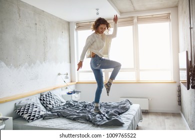 Young Happy Woman Jumping On Bed In Morning, Stylish Bedroom, Positive Mood, Wearing Jeans And Knitted White Sweater, Cheerful, Active Life, Energy, Having Fun, Slim Body, Smiling, Happy