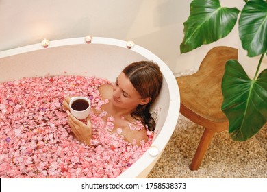 Young Happy Woman Lie In Bath Tub Full Of Pink Roses Flower Petals Drink Herbal Tea, Enjoy Her Day In The Spa. White Bath Tube With Pink  Tropical Flowers Petals, Spa, Relaxation, Body Care, Therapy