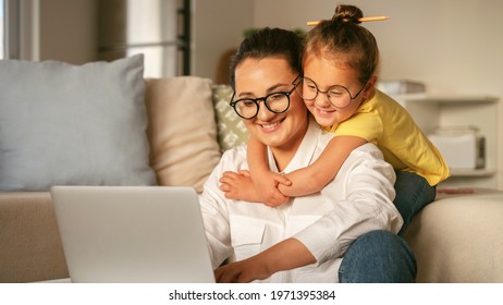 Young Happy Woman Mother In Glasses Working On Laptop With Little Girl Daughter Wearing Glasses, Excited Child Hugging Smiling Mom While Learning Online Together In Light Cozy Living Room At Home