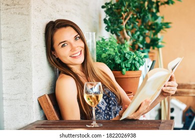 Young Happy Woman Relaxing On The Balcony, Reading Book, Leaning On The Wall, Drinking Glass Of Wine