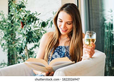 Young Happy Woman Relaxing On The Balcony, Reading Book, Holding Glass Of Wine