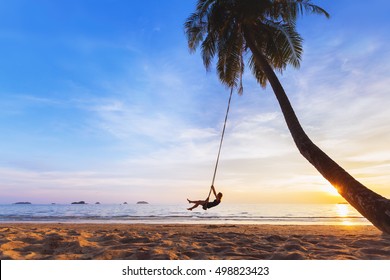 Young happy woman relaxing on a swing attached to a palm tree on a paradise beach at sunset while on vacation in a tropical country - Powered by Shutterstock