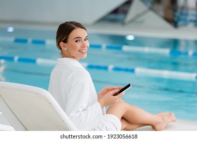 young and happy woman resting on deck chair while holding smartphone near pool