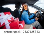 Young happy woman in Santa hat driving car with Christmas gift boxes nearby, smiling at camera, enjoy winter holiday preparation