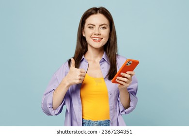 Young happy woman she wear purple shirt yellow t-shirt casual clothes hold in hand use mobile cell phone show thumb up isolated on plain pastel light blue background studio portrait. Lifestyle concept - Shutterstock ID 2367234351