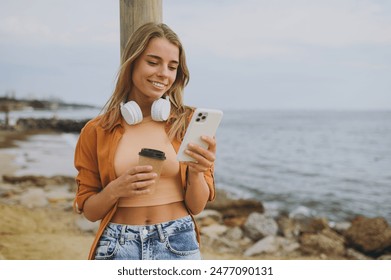 Young happy woman she wear orange shirt casual clothes headphones listen to music use mobile cell phone drink coffee walk on sea ocean sand shore beach outdoor seaside in summer day. Lifestyle concept - Powered by Shutterstock