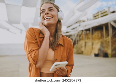 Young happy woman she wears orange shirt beige top casual clothes listen to music in headphones use mobile cell phone look aside rest relax walk outside in summer day. Urban lifestyle leisure concept - Powered by Shutterstock