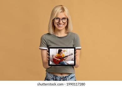 Young Happy Woman Showing Tablet Pc With Online Guitar Lesson On Screen Over Beige Studio Background. Millennial Lady Taking Remote Music Class, Using Modern Technologies. Distance Education