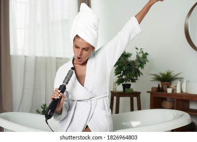 Young Happy Woman Sits On The Edge Of The Bathroom And Sings Into A Hair Dryer. Funny Woman Singing In Hairdryer At Home