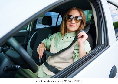 Young Happy Woman Sits At The Wheel Of A Car And Hold Seat Belt Looks At Window. Side View