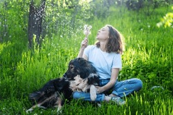 Young Happy Woman Sitting On The Grass With Her Dog Blowing On Dandelions On A Summer Day