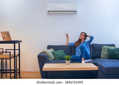 Young Happy Woman Sitting On Couch Under Air Conditioner And Adjusting Comfort Temperature With Remote Control At Modern Home