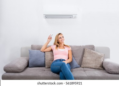 Young Happy Woman Sitting On Couch Operating Air Conditioner With Remote Control At Home