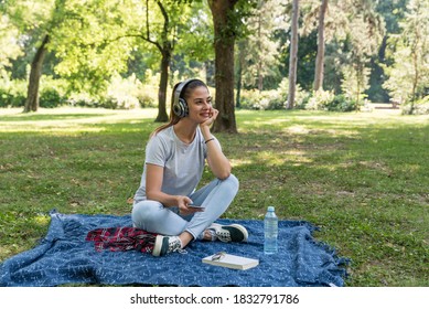 Young Happy Woman Sitting In The Park Surfing The Song Play List In Her Phone And Listening The Music On The Wireless Headphones